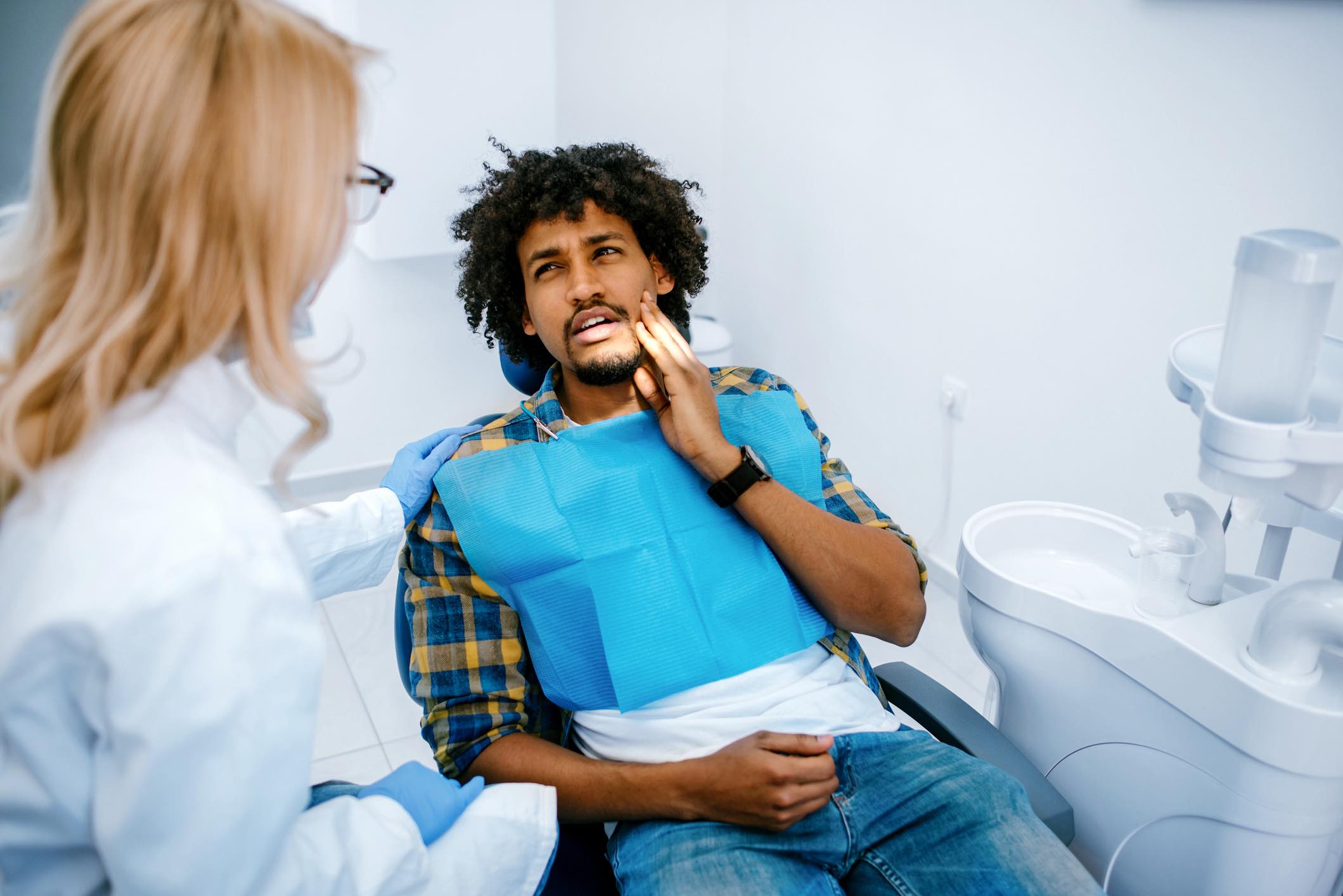 Young black man holding cheek in chair at dentist, having toothache.